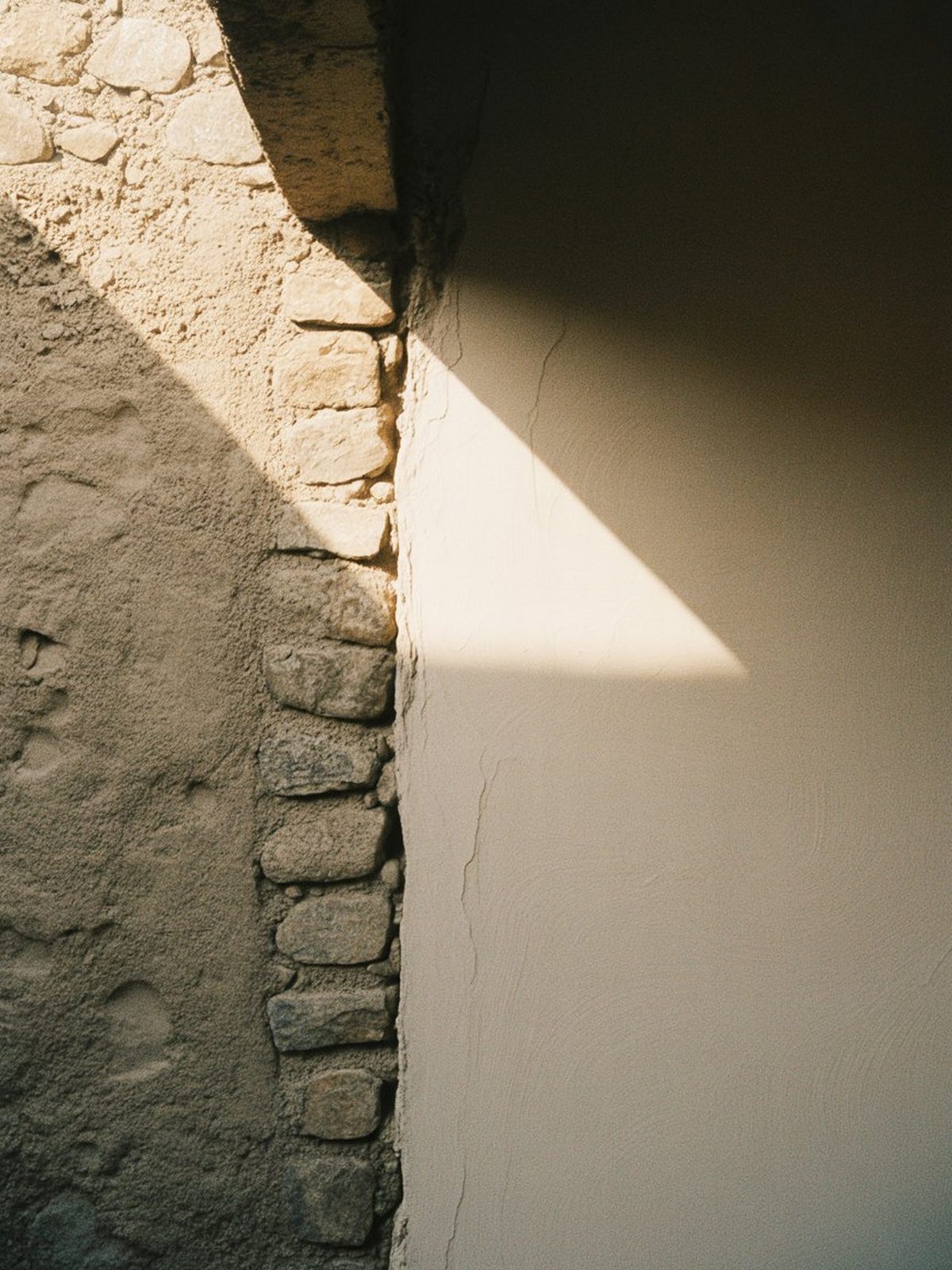 Rough stone wall meeting smooth plaster, divided by a hard diagonal of warm light