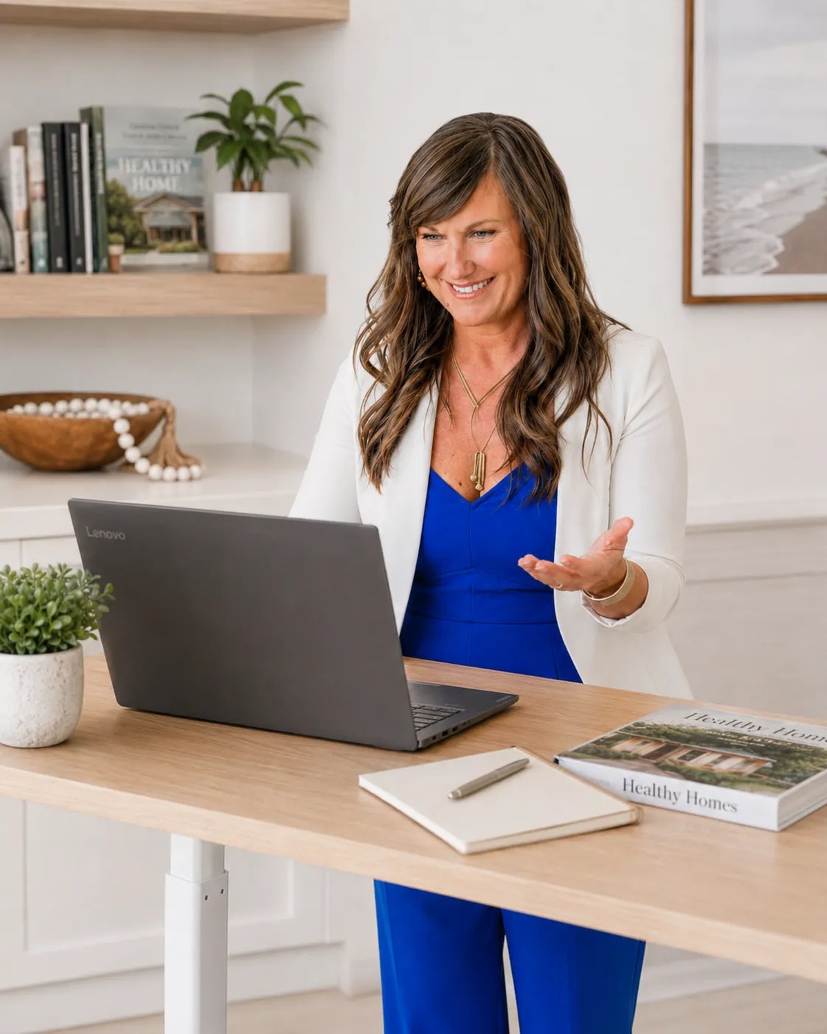 Dr. Brooke Breitbach standing at a laptop in the same blue jumpsuit and white blazer, healthy-home reference books visible on shelf, warm gesture