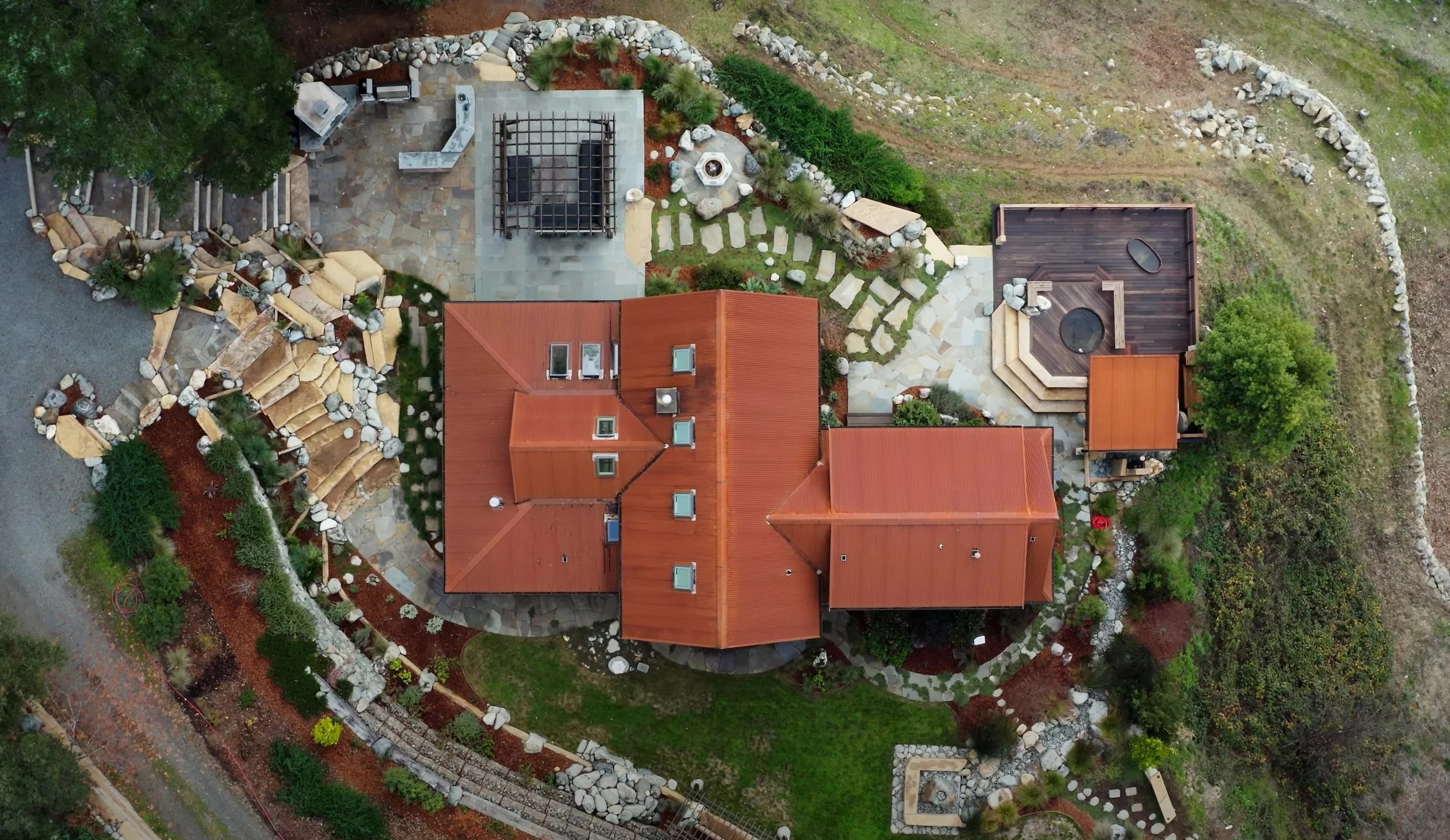 Aerial view of a terracotta-roofed timber-frame home with outdoor patio, fire pit, hot tub deck, and natural rock landscaping