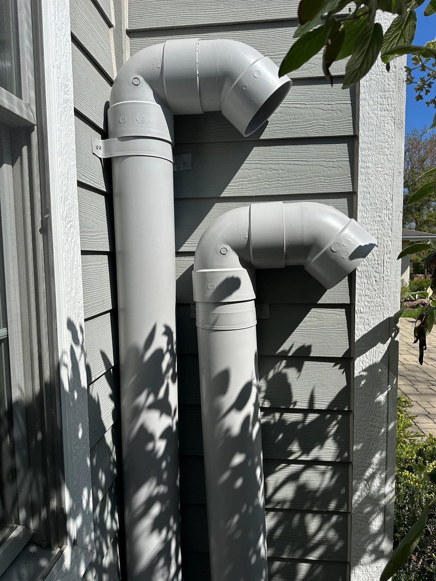 Twin gray PVC ventilation stubs at exterior corner with dappled leaf shadows