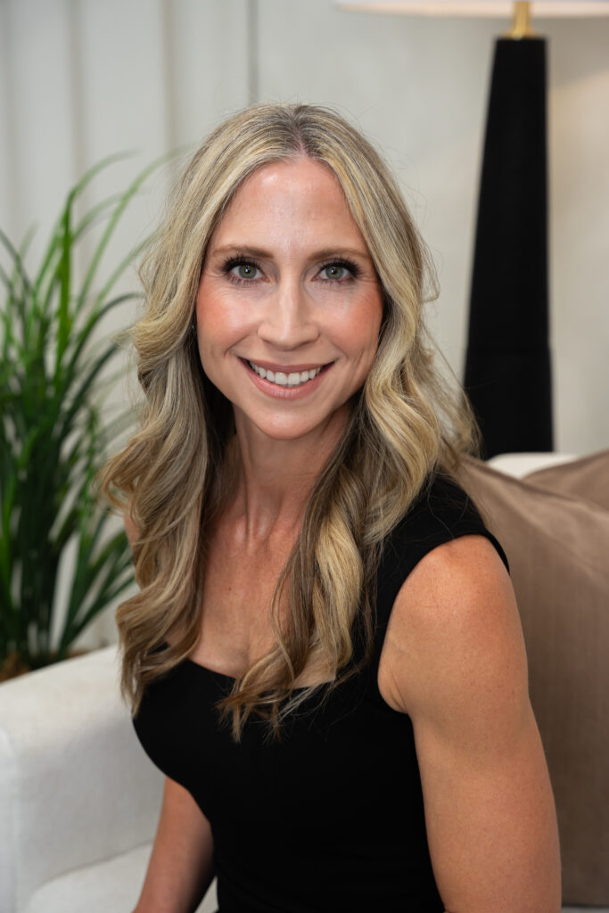 Suzanne Volkman — professional studio portrait, warm smile, seated in a styled living room with a tropical plant and lamp